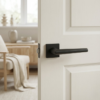 Jasmina minimalist square internal door handles in matt black, displayed on a white internal door with clean panel detailing and a soft, neutral living room backdrop.
