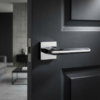 Minimalist square internal door handles in polished chrome, showing the Jasmina lever on a square rosette fitted to a dark interior door with a contemporary living space in the background.